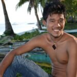 Young Filipino community leader Miguel Santos sitting by coastal area with traditional bancas, representing CJF Philippines Youth for Resilience program